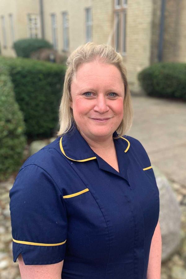 A woman with blond shoulder length hair wearing a navy nurses uniform with yellow piping on the collar, pockets and sleeves. She is smiling warmly
