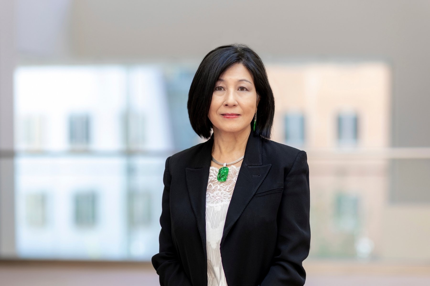 A woman with black shoulder length hair, wearing a smart black jacket, lacy white top underneath and a necklace with a jade pendant