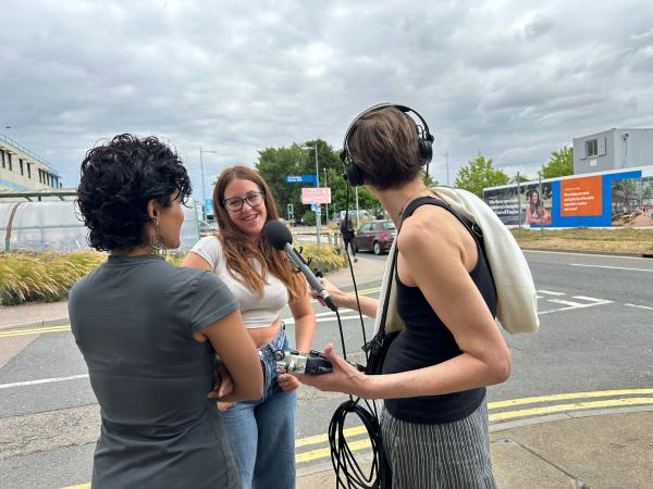 A teenage girl with long brown hair being interviewed by two women with a microphone
