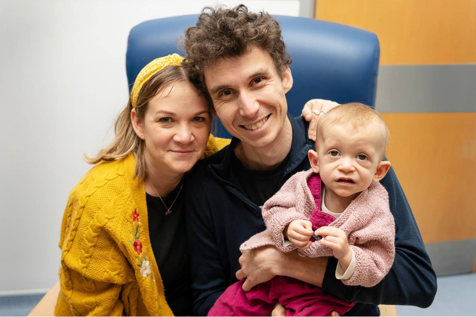 A woman with dark blond hair and a yellow hairband, with a yellow flowery cardigan, sitting alongside a man with dark curly hair and blue jumper. He is holding a baby in a pink cardigan and dungarees on his lap