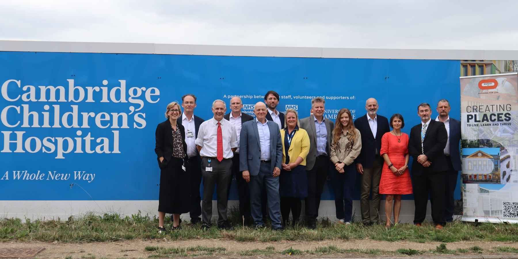 A photo of a group of people, men and women, standing in front of the bright blue Cambridge Children's Hospital hoardings by the hospital site. The hospital logo is on the left with the words A Whole New Way underneath. Most people are wearing suits. Some are wearing bright colours