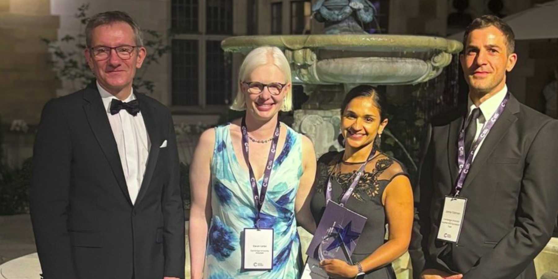 Four people in smart outfits standing by a stone fountain. The woman in the middle is holding a glass award.