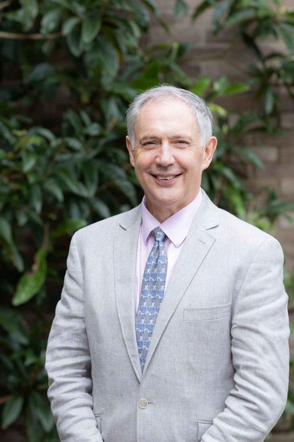A man with grey, slightly receding hair, wearing a pale grey suit, pale pink shirt and pale blue patterned tie. He is smiling. The back drop is a wall covered in dark green foliage