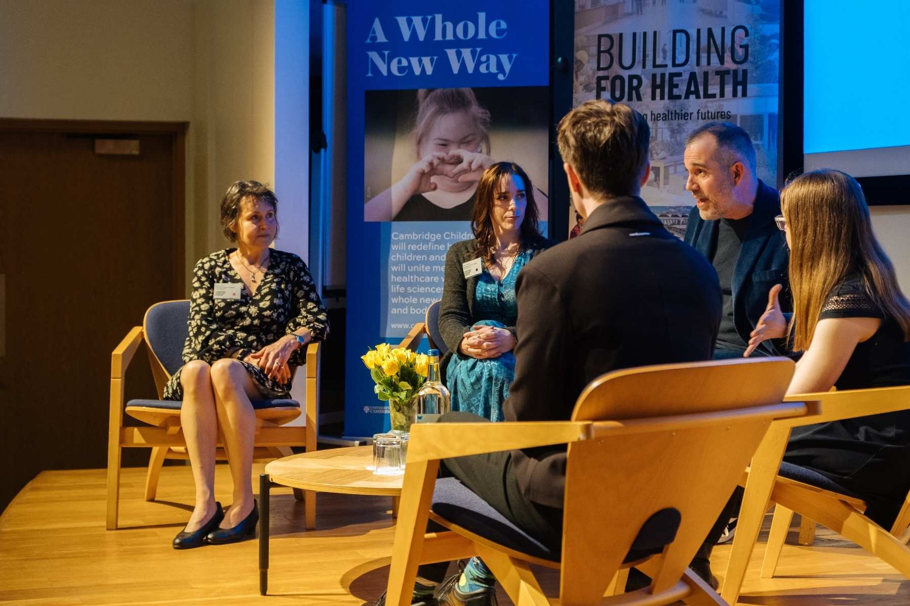 A side view image of the stage at a conference, with people sitting on low chairs around a small table with yellow roses on. A man with dark grey hair (Dr Xand) is hosting a panel discussion with the people at the children's hospital conference